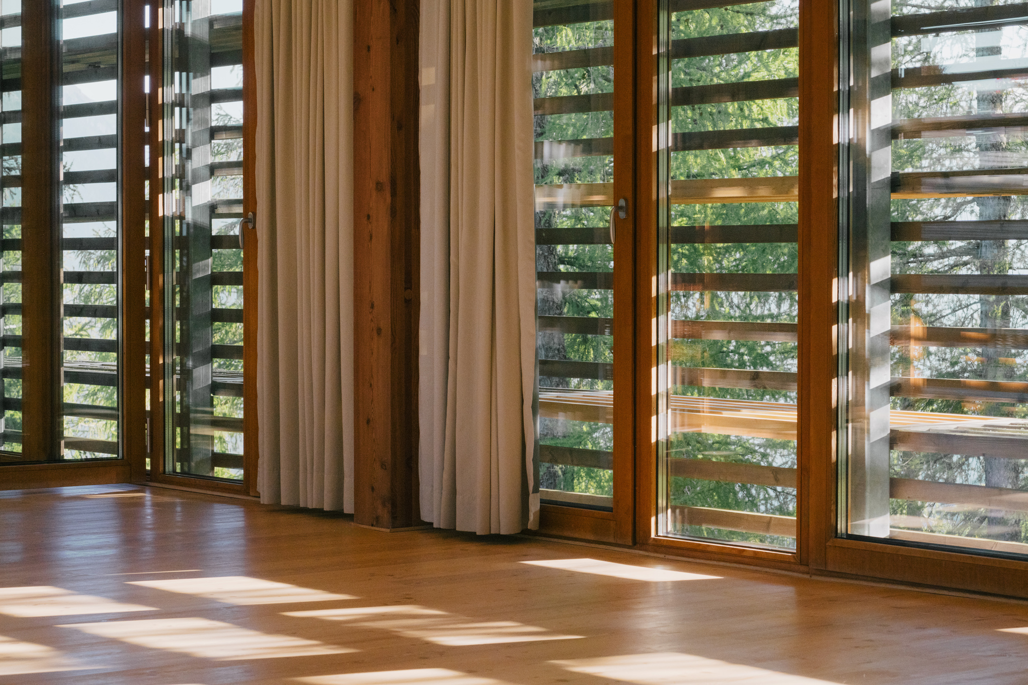 Sunlight filters through wooden slatted windows, casting shadows on a wooden floor in a spacious interior.
