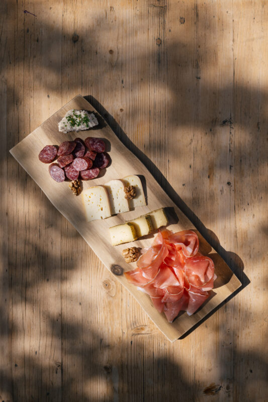 A wooden platter with assorted meats, cheeses, and a small dish of spread, set on a textured wooden surface.