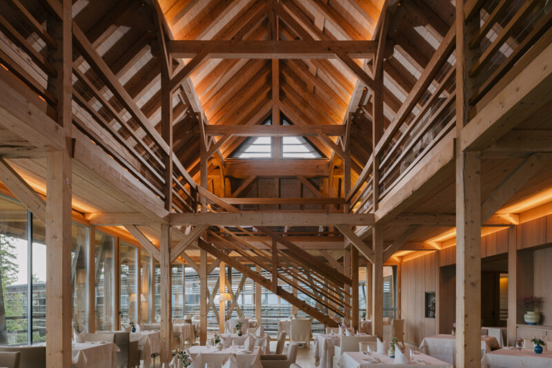 Modern wooden restaurant interior with high ceilings, large windows, and elegant table settings. Warm lighting enhances th...