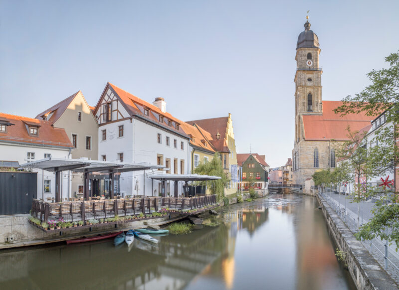 Historic buildings along a canal, with a restaurant terrace and a clock tower in the background, captured in soft evening ...
