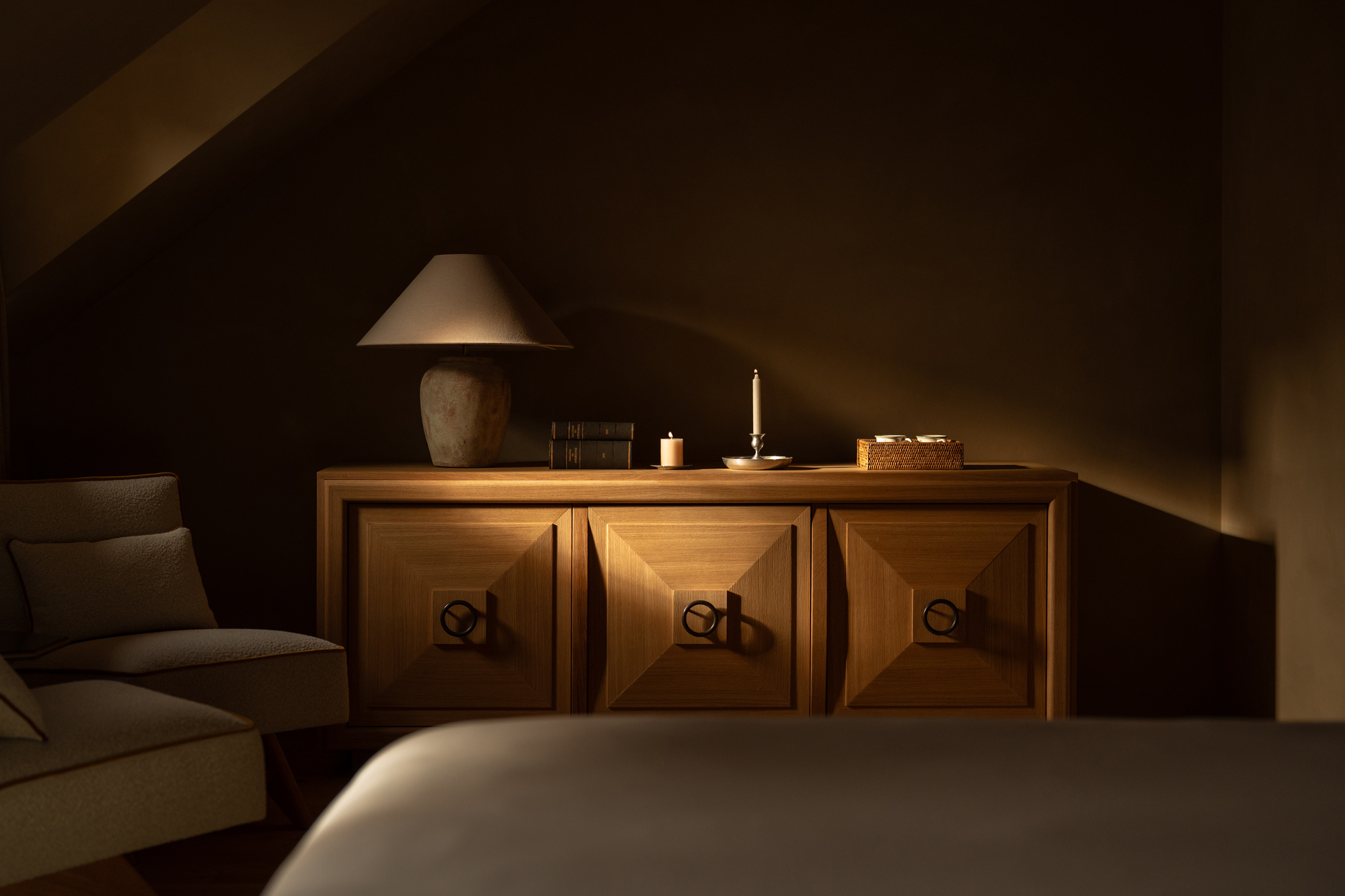 Warmly lit interior of a room featuring a wooden sideboard, a lamp, and decorative items, with soft shadows and earthy tones.