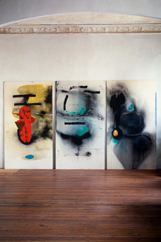 Three abstract paintings with bold colors and shapes displayed against a light wall in a wooden-floored studio.