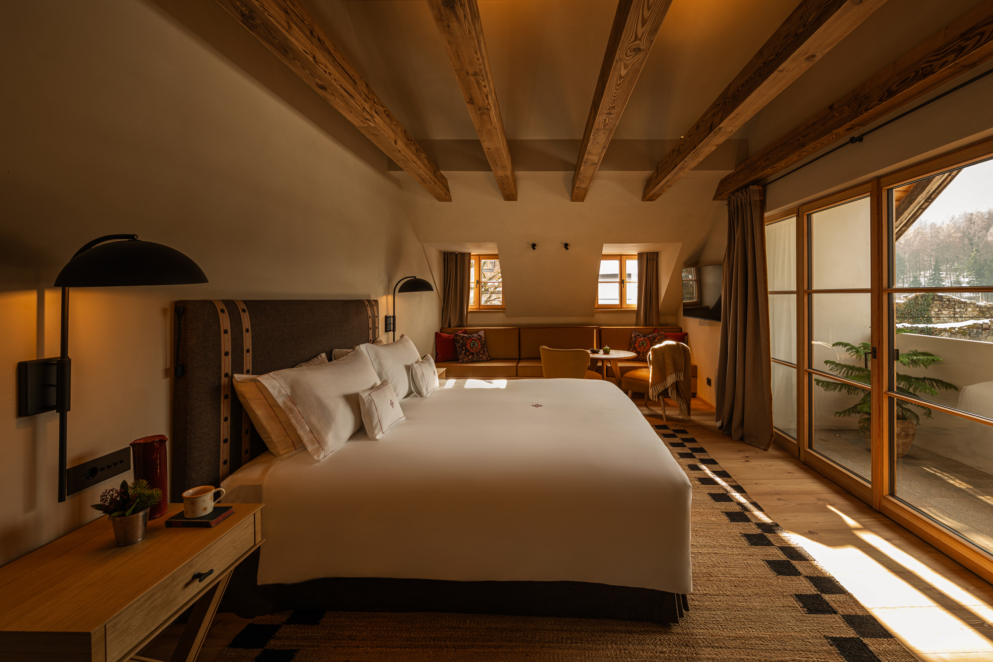 Modern hotel room at Castel Badia featuring a large bed, wooden beams, and a balcony with outdoor seating.
