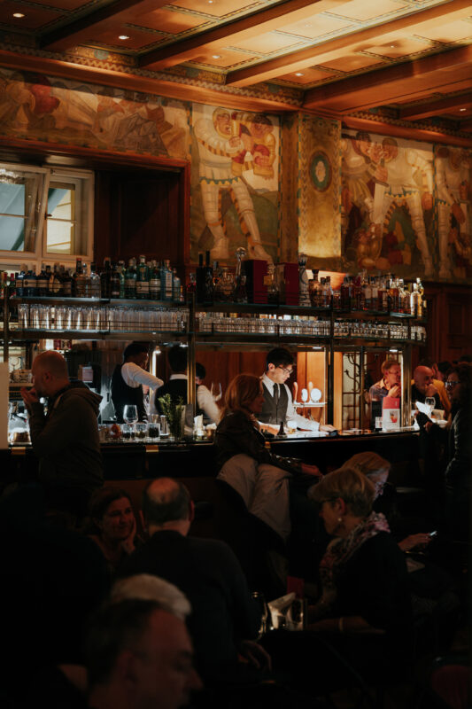 Elegant bar scene at Parkhotel Laurin, featuring patrons and bartenders amidst ornate wall murals.