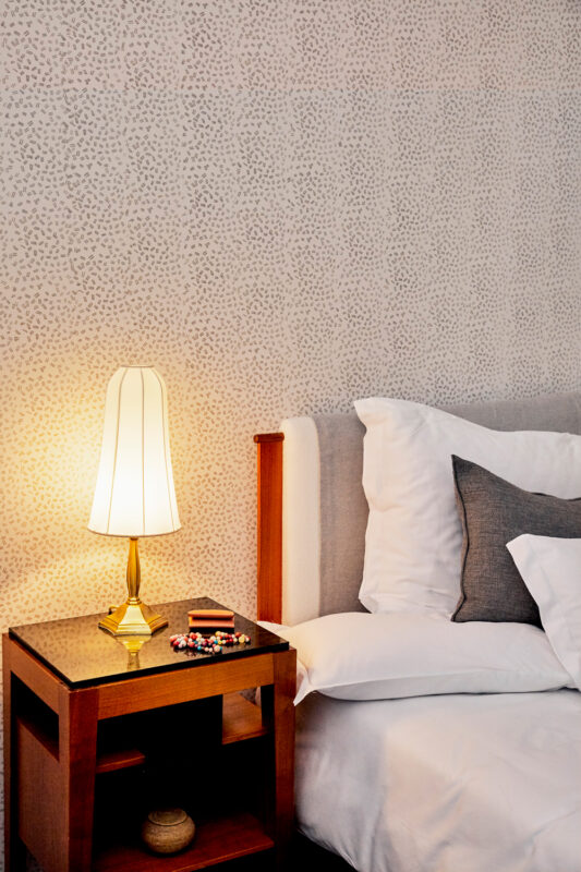 Cozy hotel room corner with a bedside table, lamp, and neatly arranged pillows against a patterned wall.
