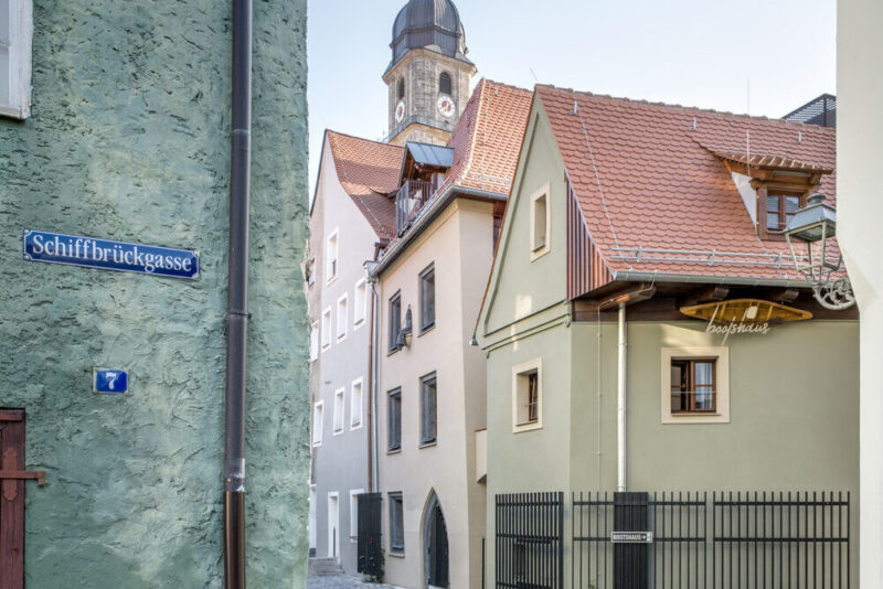 Narrow cobblestone street with buildings, including a green house and a tower in the background, near Schiffbrückgasse.