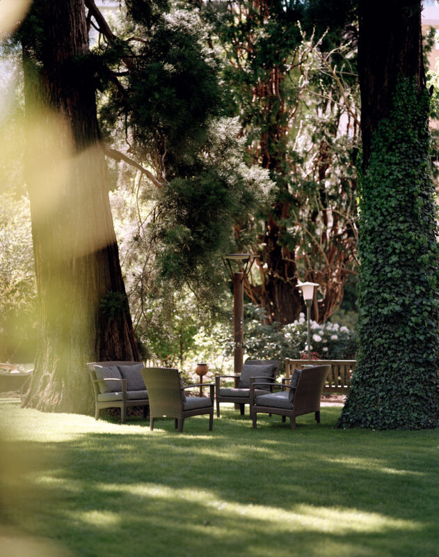 Seating area with wicker chairs and a table under tall trees in a lush green garden at Parkhotel Laurin.
