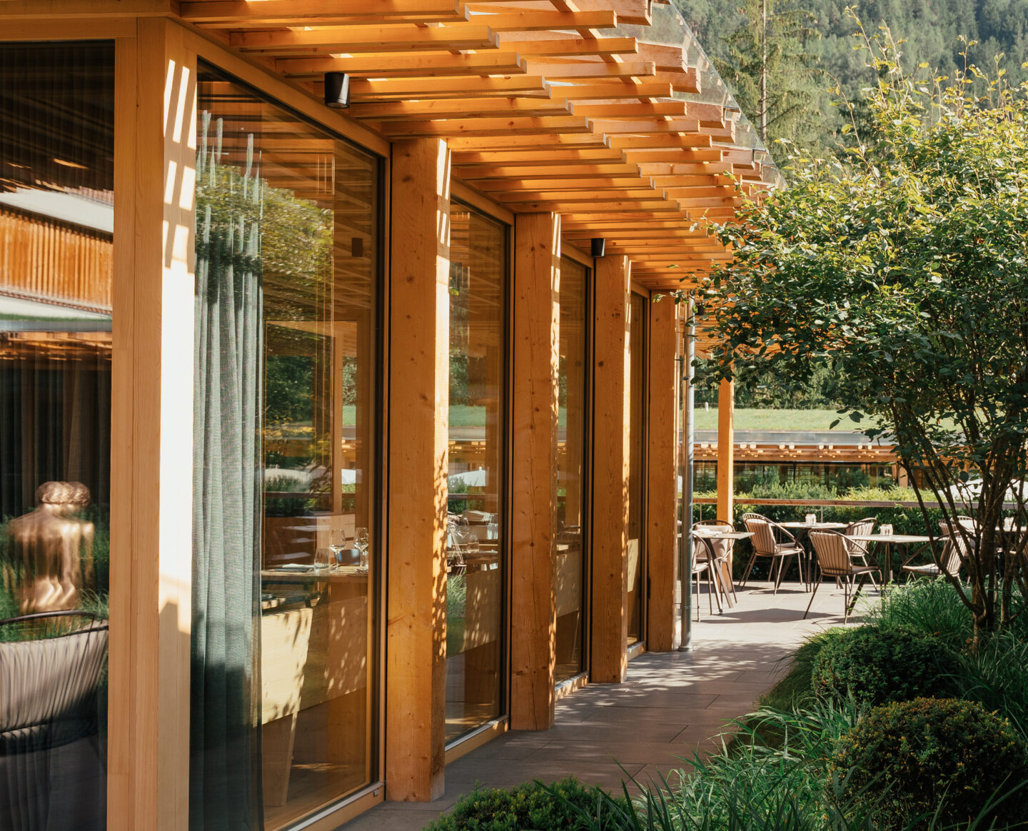 Modern wooden building with large glass windows, surrounded by greenery and outdoor dining area in the Dolomites.