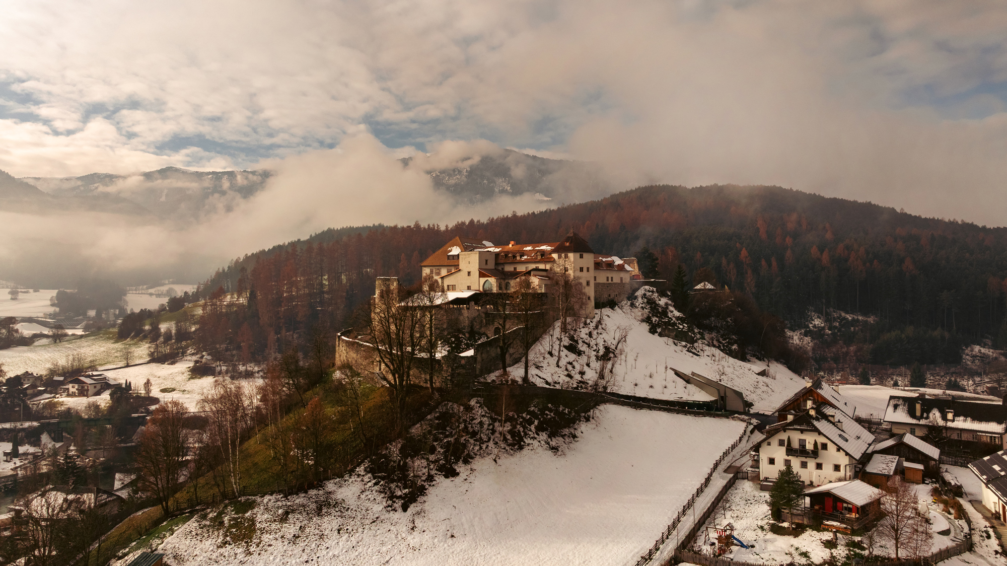 Castel Badia on a snowy hillside, surrounded by misty mountains and a forest of autumn-colored trees.