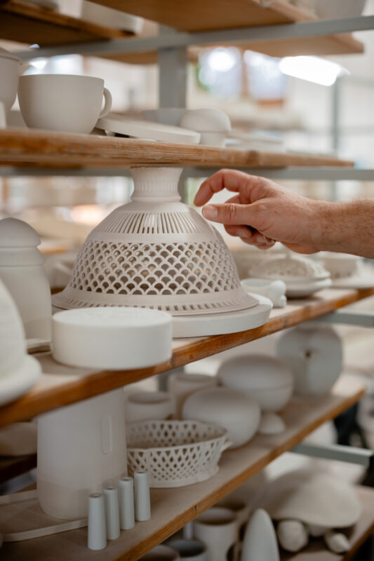 A hand reaches for a decorative white porcelain piece on a wooden shelf filled with various ceramic items.