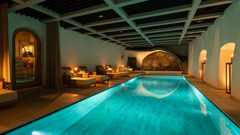 Indoor swimming pool at Castel Badia, featuring lounge chairs and ambient lighting, with a natural stone wall in the backg...