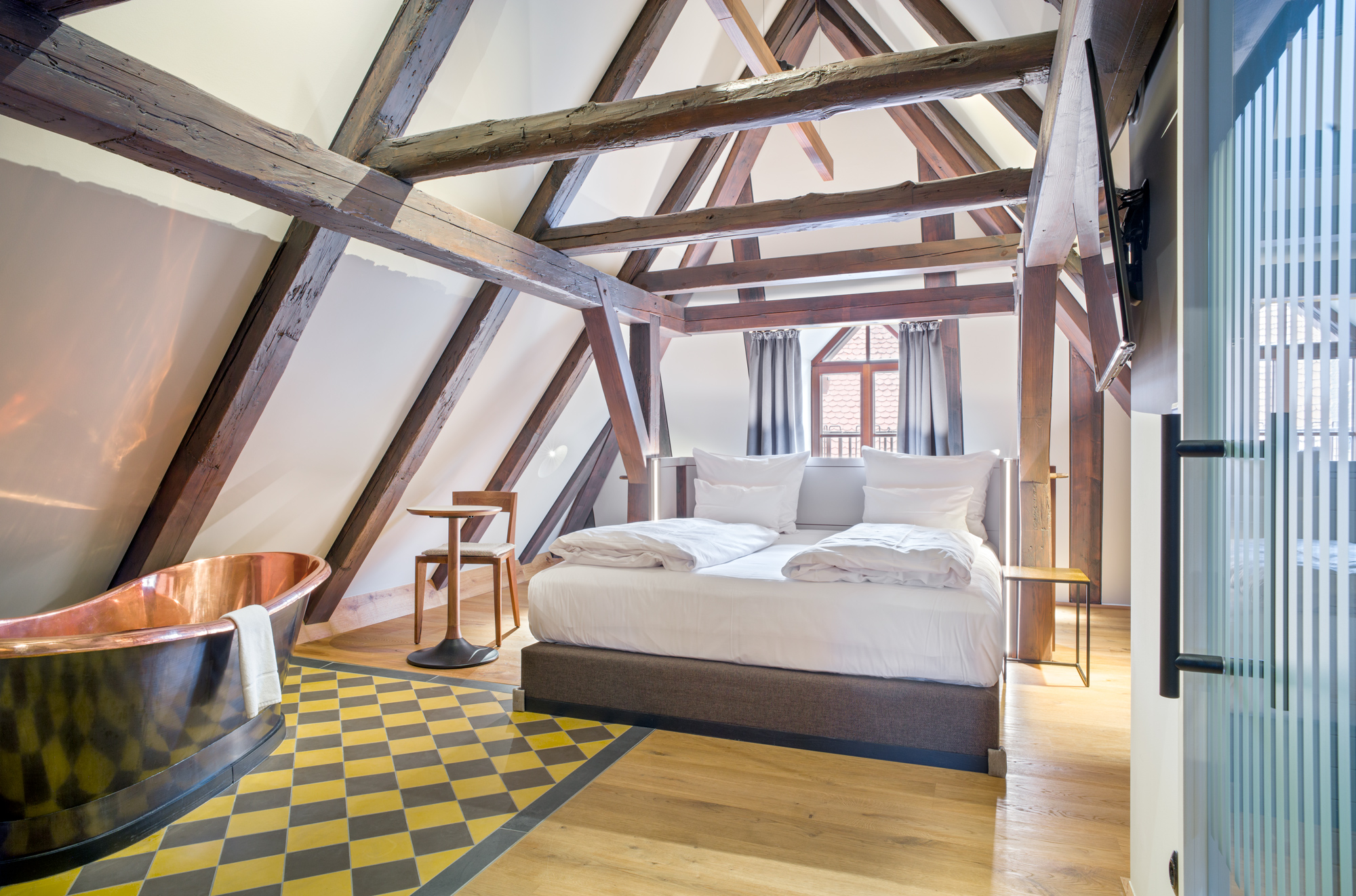 Stylish bedroom in a loft with wooden beams, a copper bathtub, and a patterned floor, featured in Bootshaus.