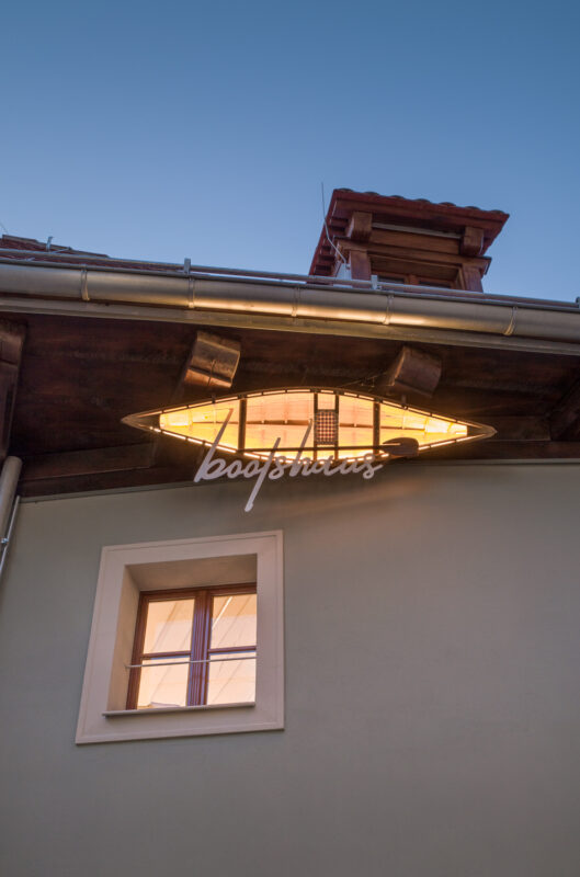 Illuminated sign reading "bootshaus" on a building under a twilight sky, with a window visible below.