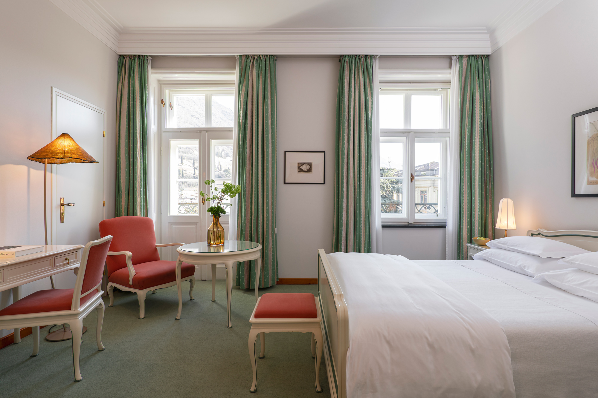Bright hotel room featuring a bed, a small table with chairs, and large windows with green curtains.