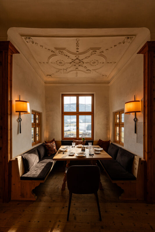 Cozy dining area in Castel Badia featuring a wooden table, upholstered benches, and large windows with scenic views.