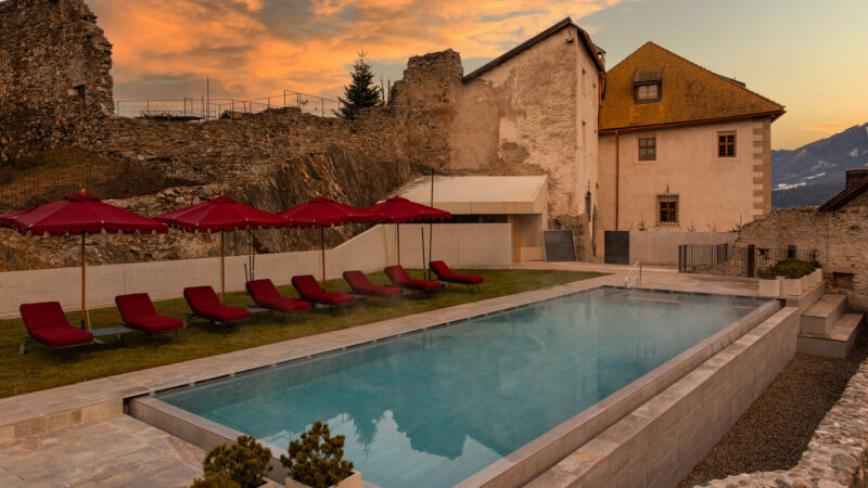 Pool area with red loungers and umbrellas, set against the backdrop of Castel Badia ruins at sunset.