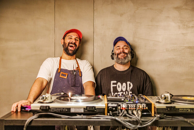 Two DJs smiling behind a turntable setup, one wearing an apron and the other in a black "FOOD" shirt.
