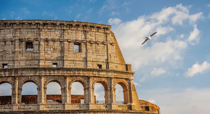 Colosseum in Rome with a bird flying in the blue sky and scattered clouds.