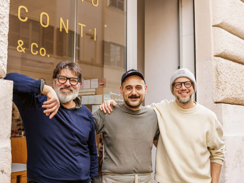 Three men stand outside a shop with a sign reading "CONTÌ & Co." in Rome, smiling and posing together.