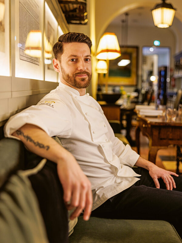 Chef sitting on a couch in a restaurant, wearing a white chef's jacket, with warm lighting and decor in the background.