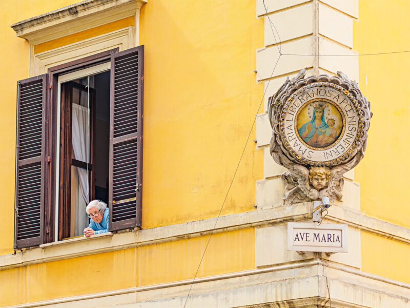 Elderly person in a blue shirt leaning out of a window on a yellow building, with a decorative plaque reading "AVE MARIA.