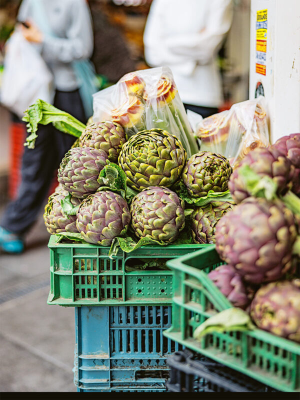 Artichokes stacked in green crates at a market, with blurred figures in the background.