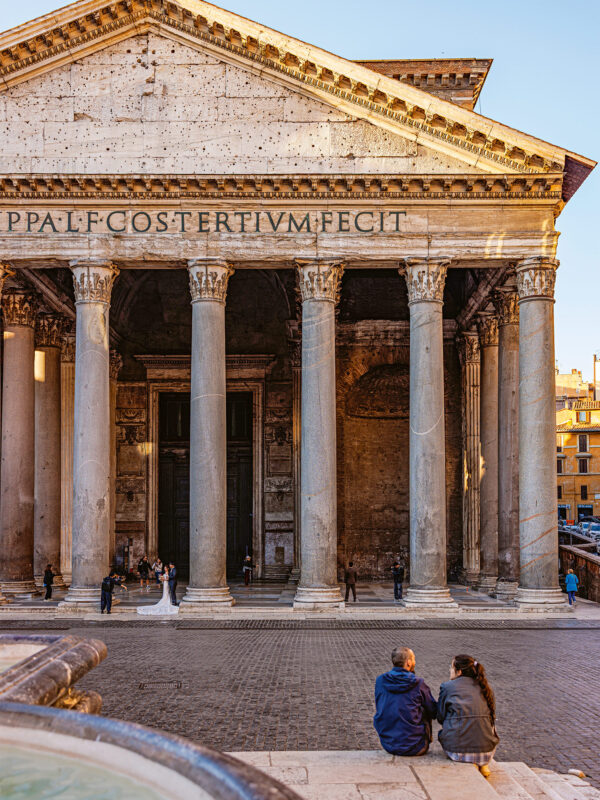 Couple sitting on steps in front of the Pantheon in Rome, with its grand columns and ancient architecture.