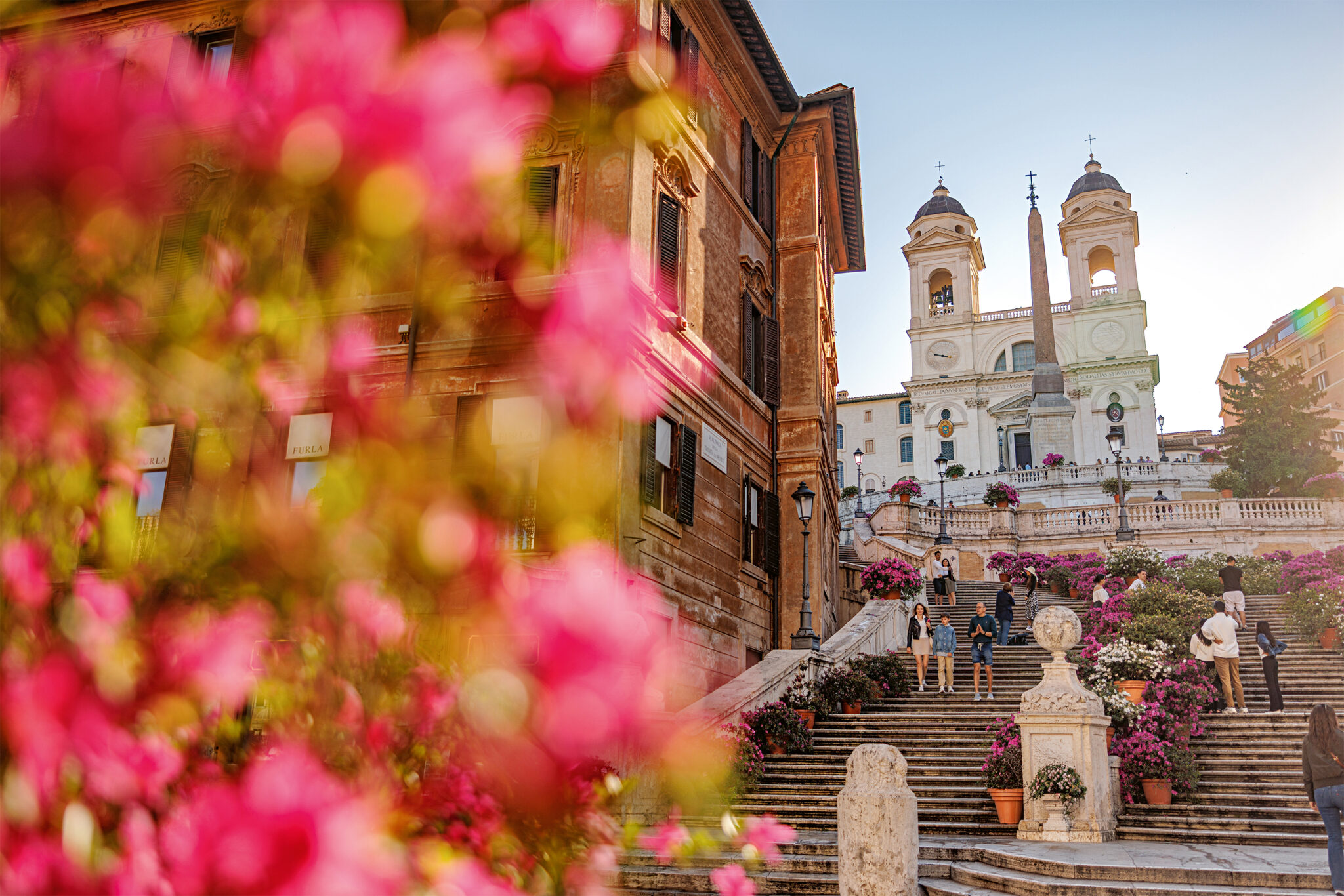 Colorful flowers in the foreground with the Spanish Steps and church in the background, bathed in warm sunlight.