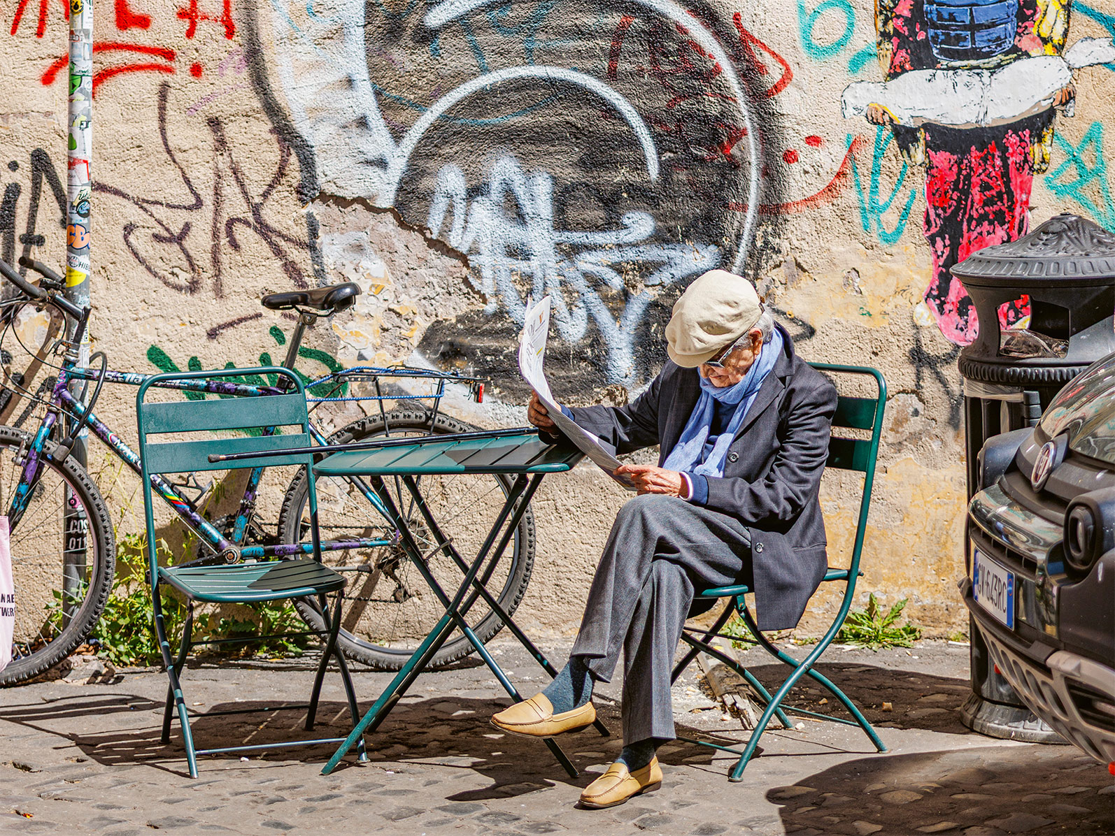 Elderly man reading a newspaper while seated at a green table near a graffiti-covered wall in Rome.