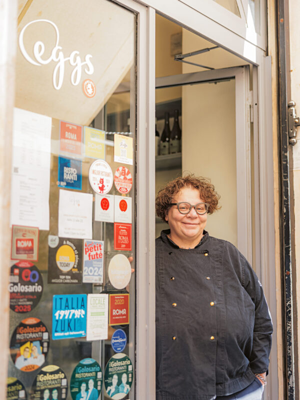 Chef standing at the entrance of a restaurant named "Eggs," surrounded by colorful stickers and signs.