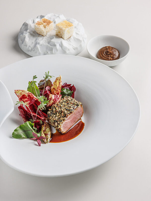 Seared meat coated in seeds, garnished with greens and sauce, with a dessert and sauce in the background.