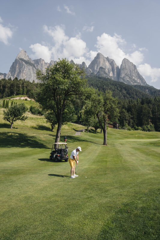 Golfer in yellow shorts preparing to swing on a lush green course, with mountains in the background.