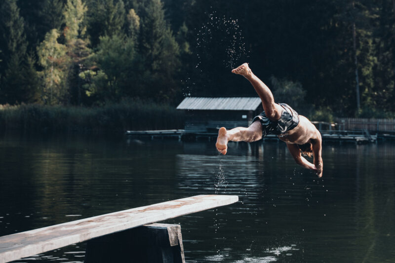 Man diving into a lake from a wooden platform, surrounded by trees and a rustic cabin in the background.