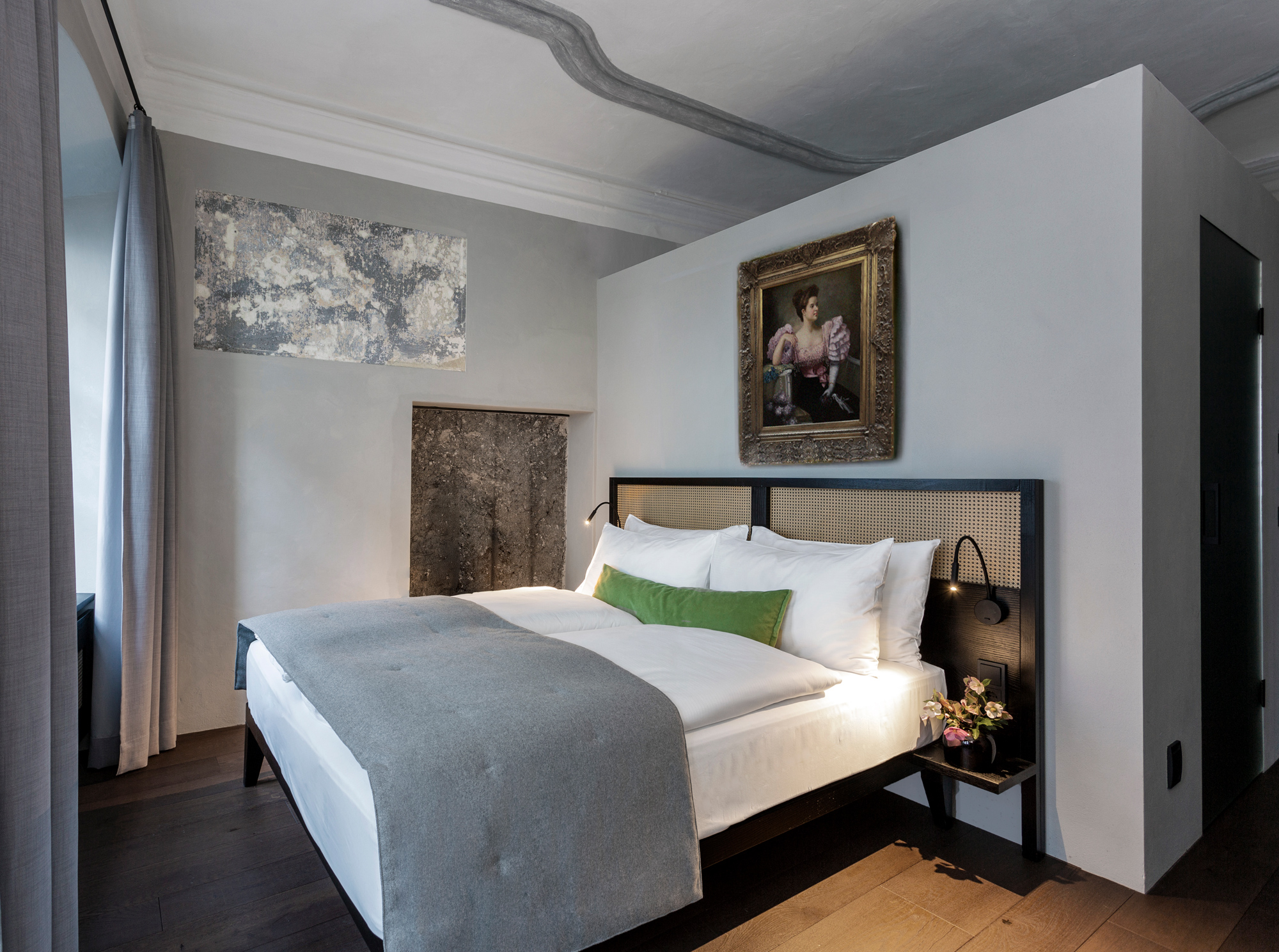 Modern hotel room featuring a double bed with gray blanket, green accent pillow, and a vintage painting on the wall.