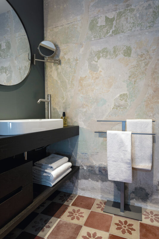 Modern bathroom featuring a round mirror, sink, towel rack with white towels, and textured wall with decorative tiles.