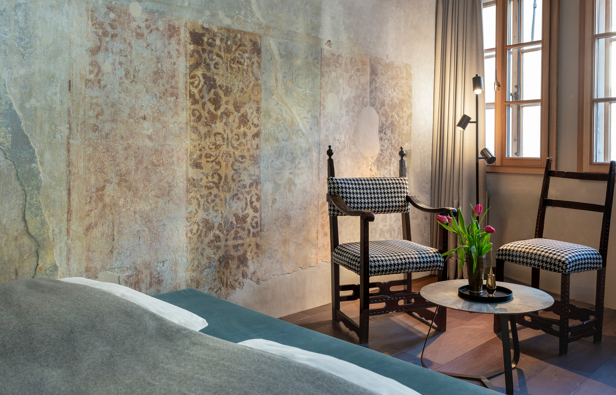 Detail of a room featuring vintage chairs, a small table with tulips, and textured wall art in soft lighting.