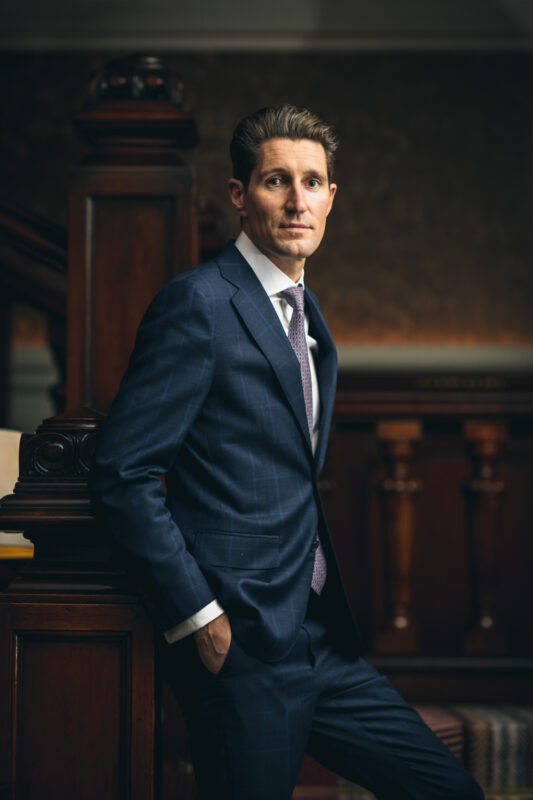 Man in a tailored blue suit leaning against a wooden railing, with a formal setting in the background.
