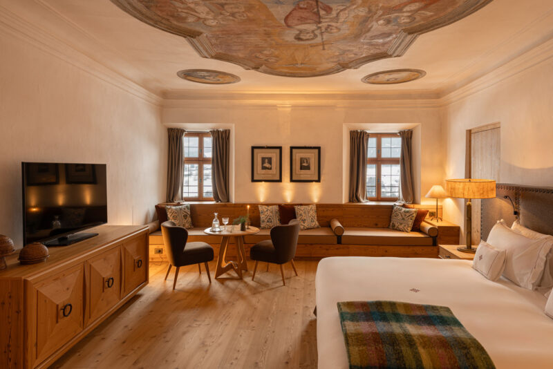 Elegant hotel room at Castel Badia featuring a frescoed ceiling, wooden furnishings, and a cozy seating area.