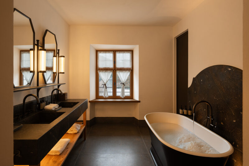 Modern bathroom featuring a freestanding bathtub, dual sinks with black countertops, and a window with sheer curtains.