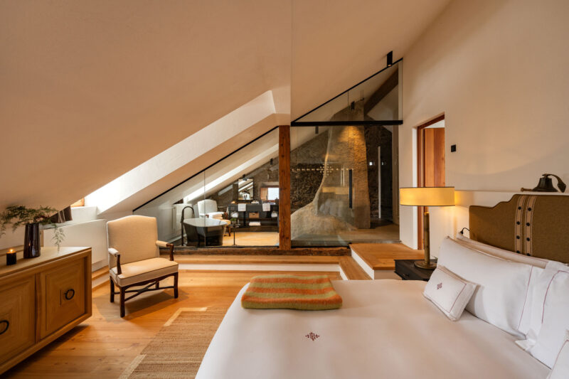 Cozy chalet bedroom with a sloped ceiling, wooden furnishings, and a view of a modern sitting area through glass walls.