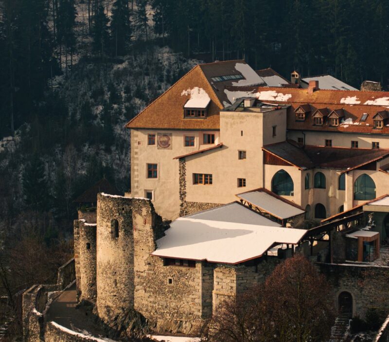 Castel Badia, a historic castle with snow-covered roofs, surrounded by trees and mountains.