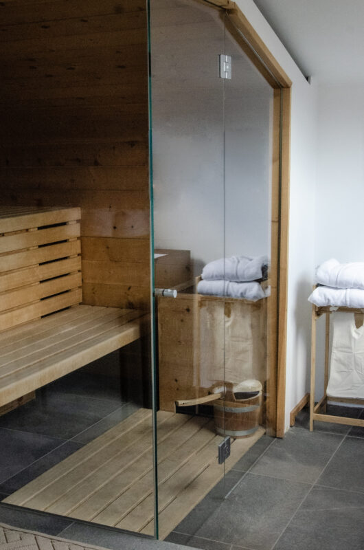Wooden sauna interior featuring a glass door, a bench, and neatly stacked towels on a wooden rack.