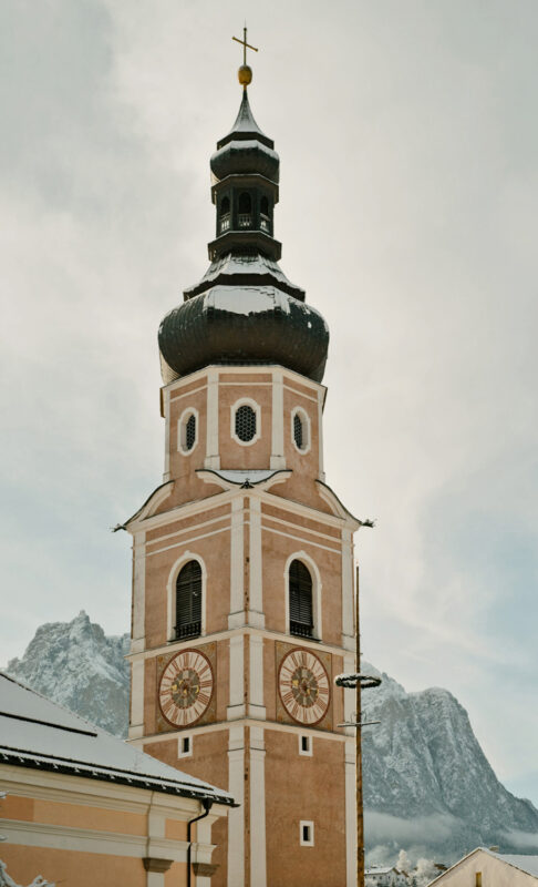 Historic church tower with a black dome and clock, surrounded by snow-covered mountains under a cloudy sky.