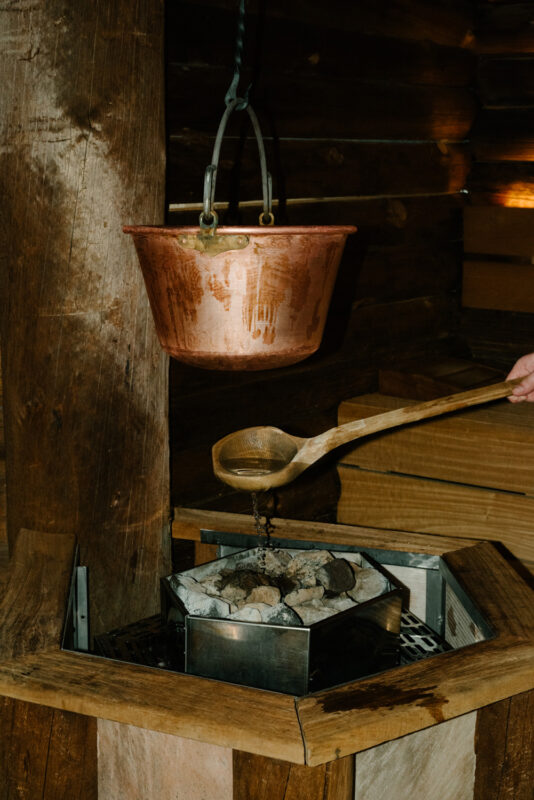 Copper pot hanging above a wooden sauna stove with a wooden ladle pouring water onto hot stones.