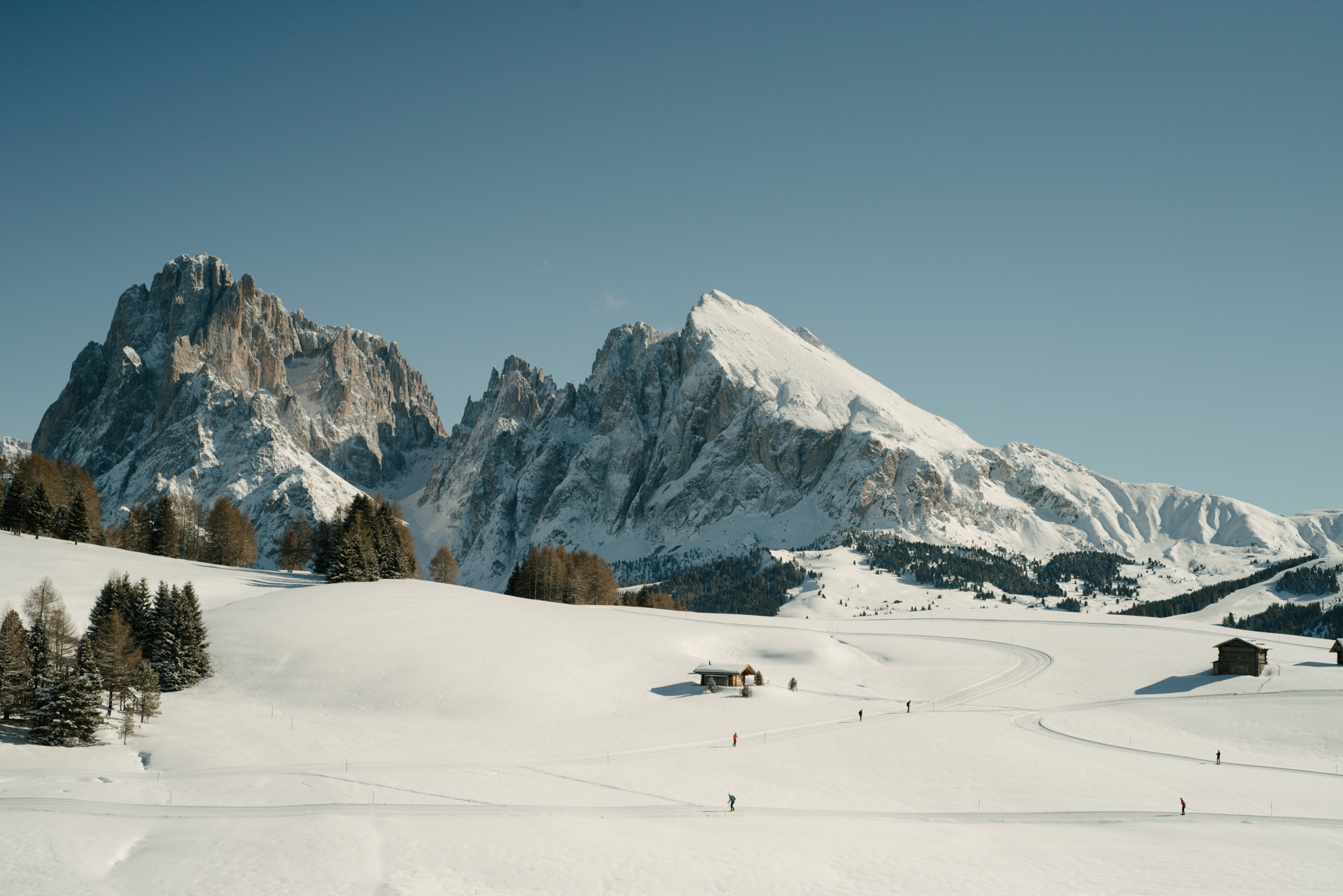Snow-covered landscape with skiers, featuring the Dolomites mountains in the background under a clear blue sky.