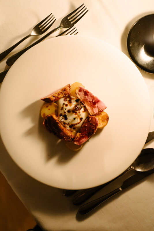 Gourmet dish featuring sliced meats and potatoes, garnished with herbs, on a white plate surrounded by cutlery.