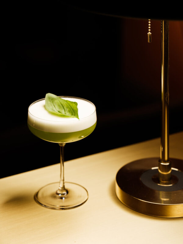 Green cocktail with a frothy top and a basil leaf garnish, placed next to a lamp on a wooden surface.