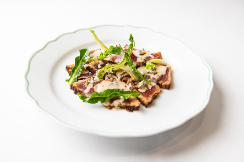 Bluefin tuna tataki served on a white plate, garnished with greens and drizzled with tonnata sauce.