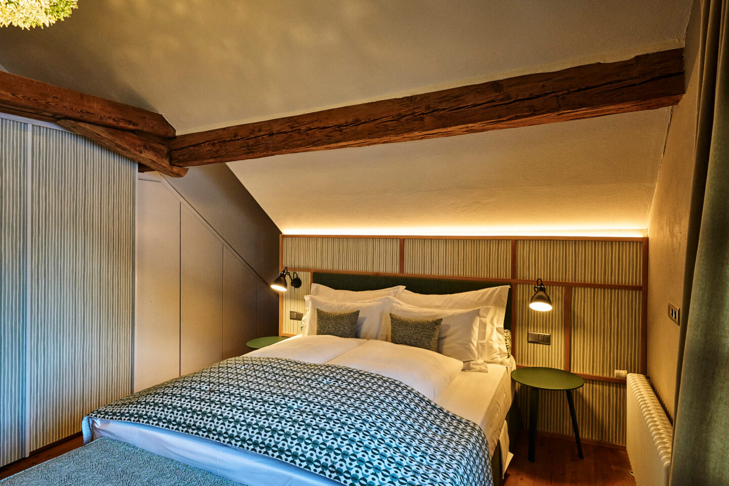 Cozy bedroom with a sloped ceiling, wooden beams, and soft lighting, featuring a double bed and bedside tables.
