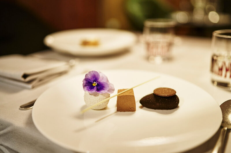 Dessert plate featuring a white mousse, chocolate disc, and a purple flower, set on a white tablecloth.